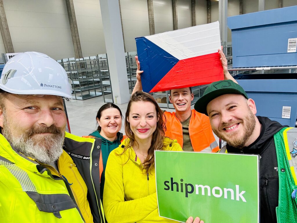 Hello, friends! Picture this: five industrious individuals standing tall and beaming with pride in a lively warehouse setting. Each is donned in essential safety gear and vibrant high-visibility clothing—a testament to the bustling energy within. The captivating scene is enhanced by one proud person holding high the Czech flag, celebrating diversity and international collaboration. Another jubilantly displays a sign emblazoned with ShipMonk, the unsung hero of third-party logistics and fulfillment solutions. At ShipMonk.com, it’s all about precision, teamwork, and making dreams come true—as if orchestrating a cinematic masterpiece where every role shines brightly together towards splendid success!. via ShipMonk.com