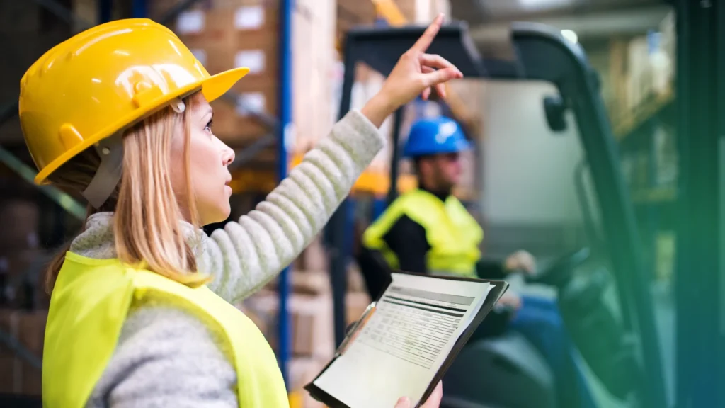 Here’s a delightful snapshot from the heart of a bustling warehouse: a woman in a cheerful yellow hard hat and bright safety vest holds a clipboard with confidence, pointing upward as she addresses what might be receiving errors with calm assurance. Nearby, another team member in blue skillfully navigates a forklift. It’s this blend of attention to detail and teamwork that keeps things running smoothly at ShipMonk.com—where tackling logistics challenges with care (and just the right dash of know-how) helps your business thrive. via ShipMonk.com