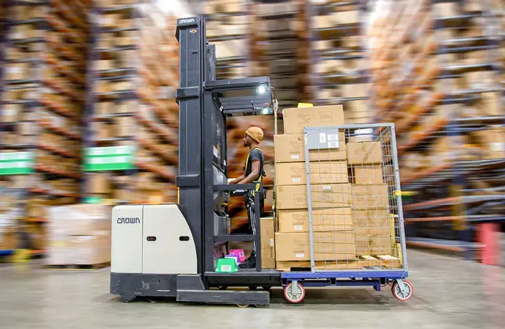 A warehouse employee expertly operates a forklift, moving a large stack of cardboard boxes secured on a metal rack. The background, featuring shelves filled with various items, creates a blurred effect typical of bustling bonded warehouses offering efficient warehousing solutions.